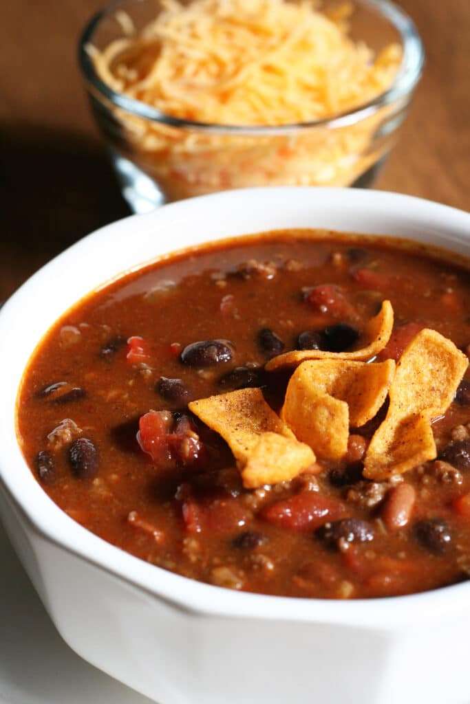 A bowl of hearty Beef & Sausage Chili filled with beans, meat, and a rich tomato base, topped with corn chips. A bowl of shredded cheese and extra chips are visible in the background