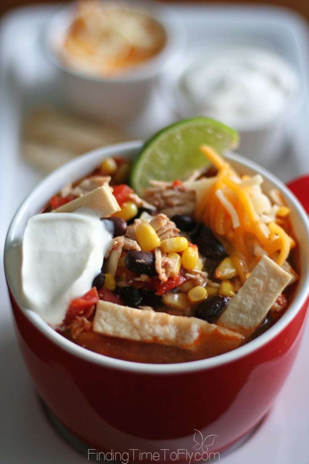 A bowl of Slow Cooker Chicken Tortilla Soup topped with crispy tortilla strips, shredded cheese, sour cream, and a lime wedge. The soup is filled with chicken, black beans, corn, and tomatoes, served in a red bowl.