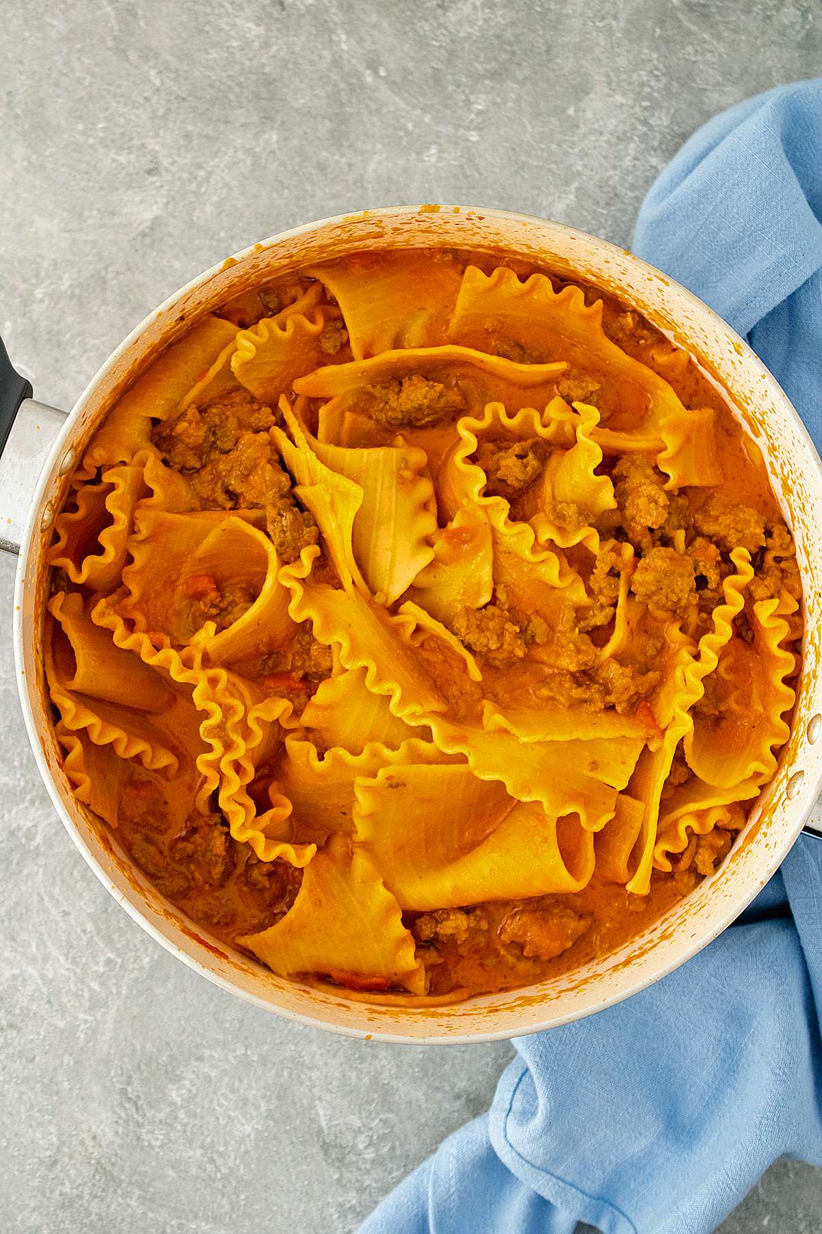 A pot of Spicy Lasagna Soup featuring wide lasagna noodles, spicy sausage, and a rich tomato broth. The dish is served in a white pot with a blue kitchen towel in the background.