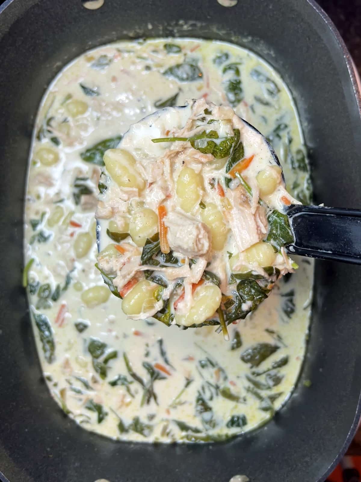 A ladle of Slow Cooker Chicken Gnocchi Soup filled with shredded chicken, gnocchi, spinach, and creamy broth, served from a slow cooker. The soup features vibrant greens and a rich, creamy texture.