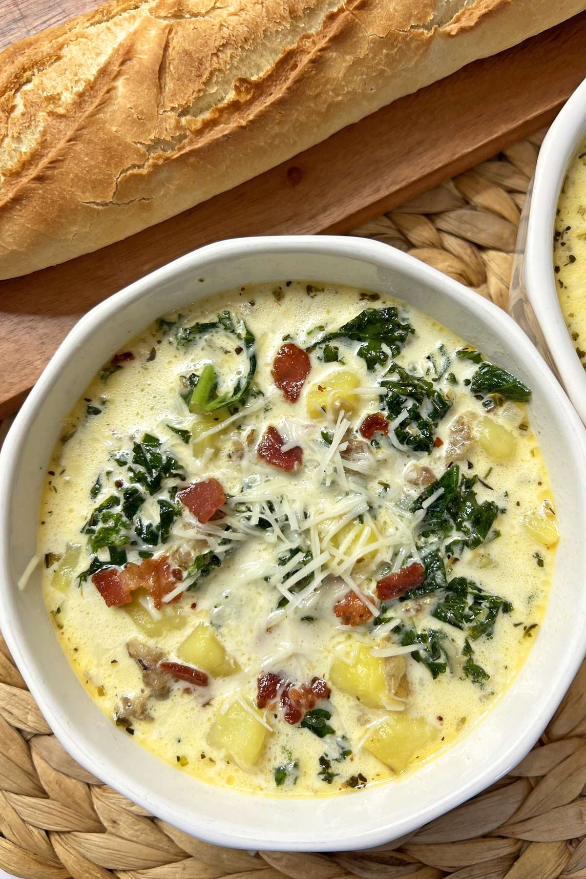 A bowl of creamy Sausage, Kale, and Potato Soup featuring chunks of potato, sausage, kale, and crispy bacon, garnished with shredded cheese. A loaf of crusty bread is visible in the background.