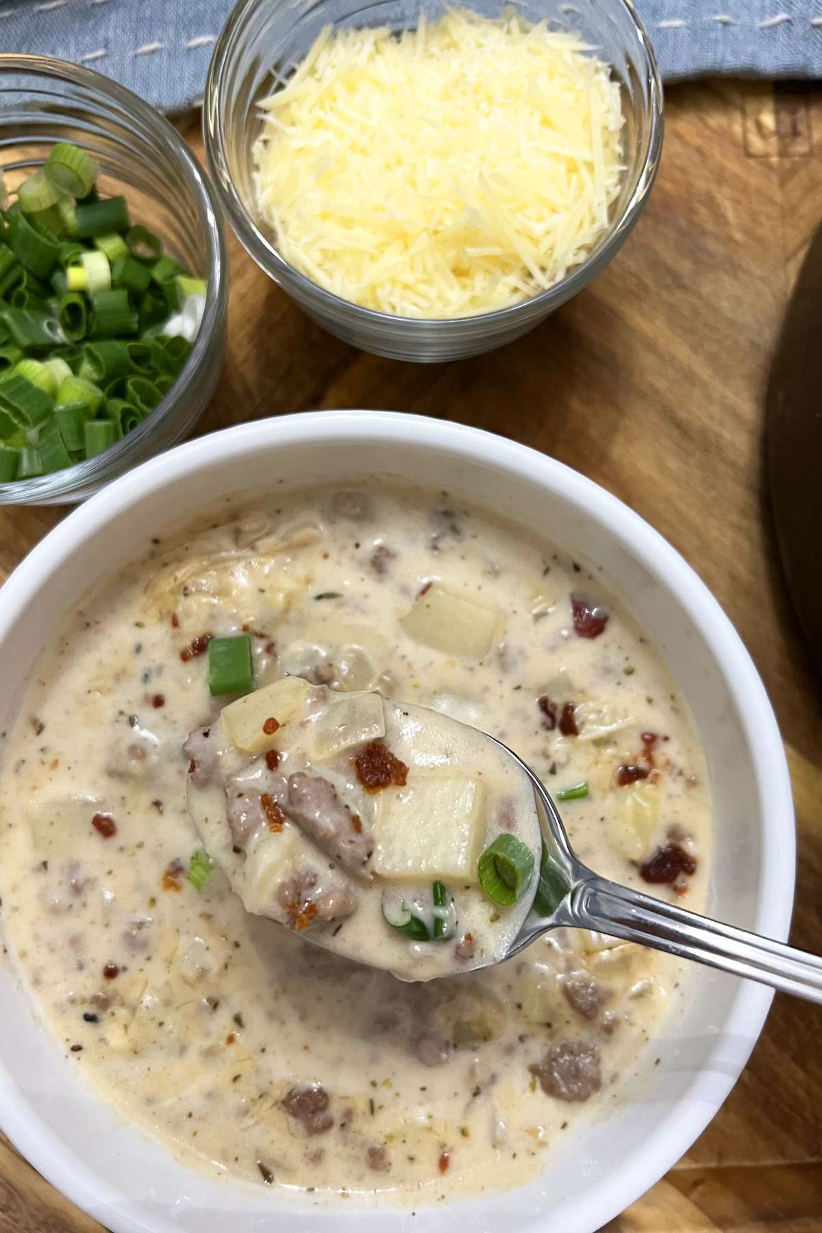 A bowl of Cheesy Hamburger Potato Soup topped with shredded cheese, crispy bacon bits, and sliced green onions, surrounded by bowls of toppings on a wooden surface.