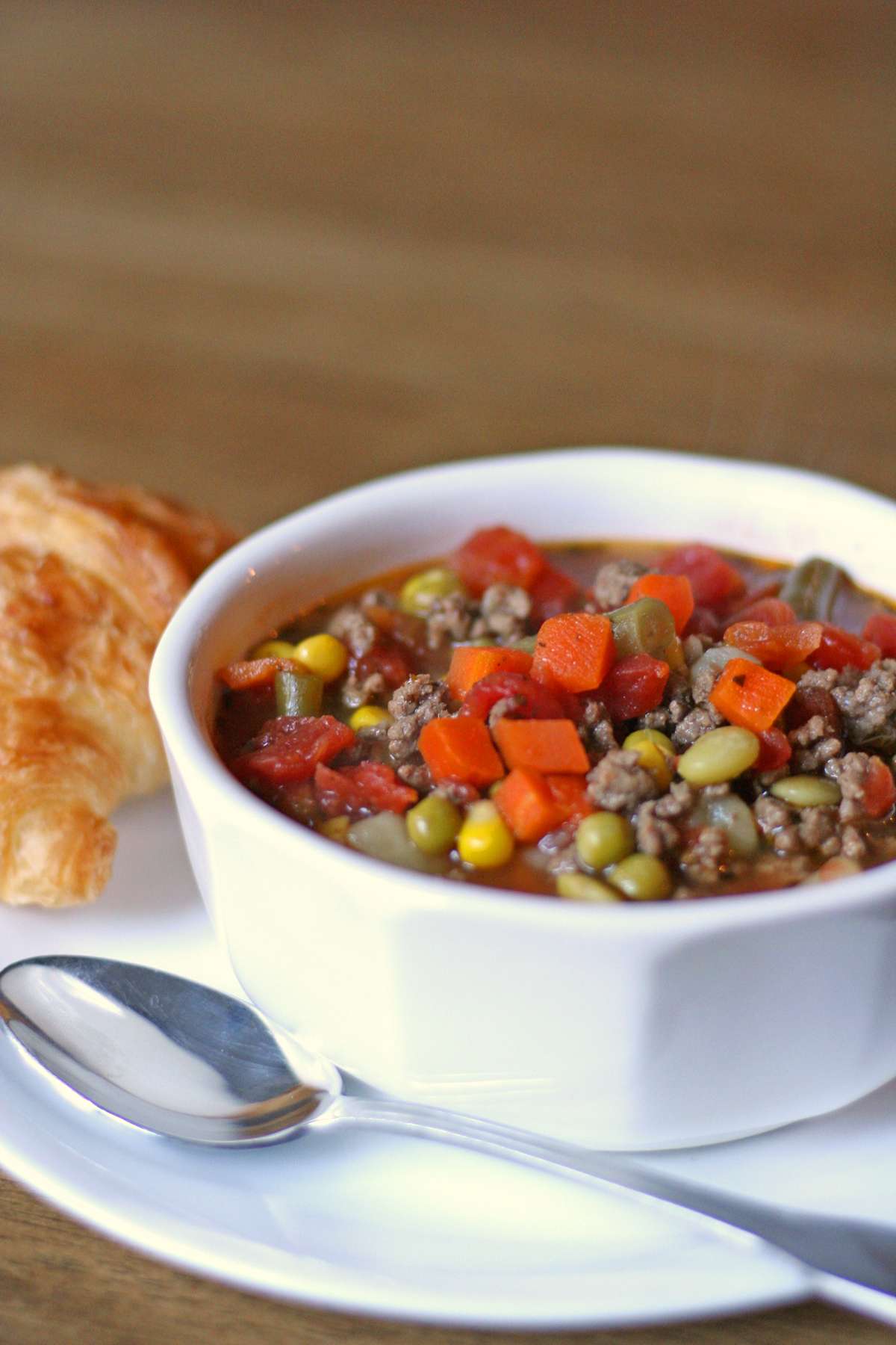 A bowl of Hamburger Vegetable Soup featuring ground beef, carrots, peas, green beans, and a tomato-based broth. Served on a white plate with a spoon and a buttery croissant on the side.