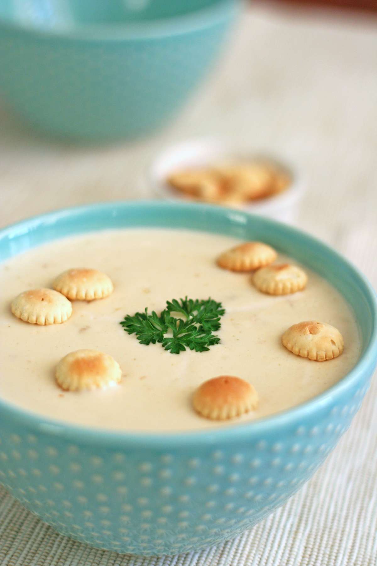 A bowl of creamy New England Clam Chowder topped with oyster crackers and a sprig of parsley, served in a blue bowl with a dish of crackers in the background.