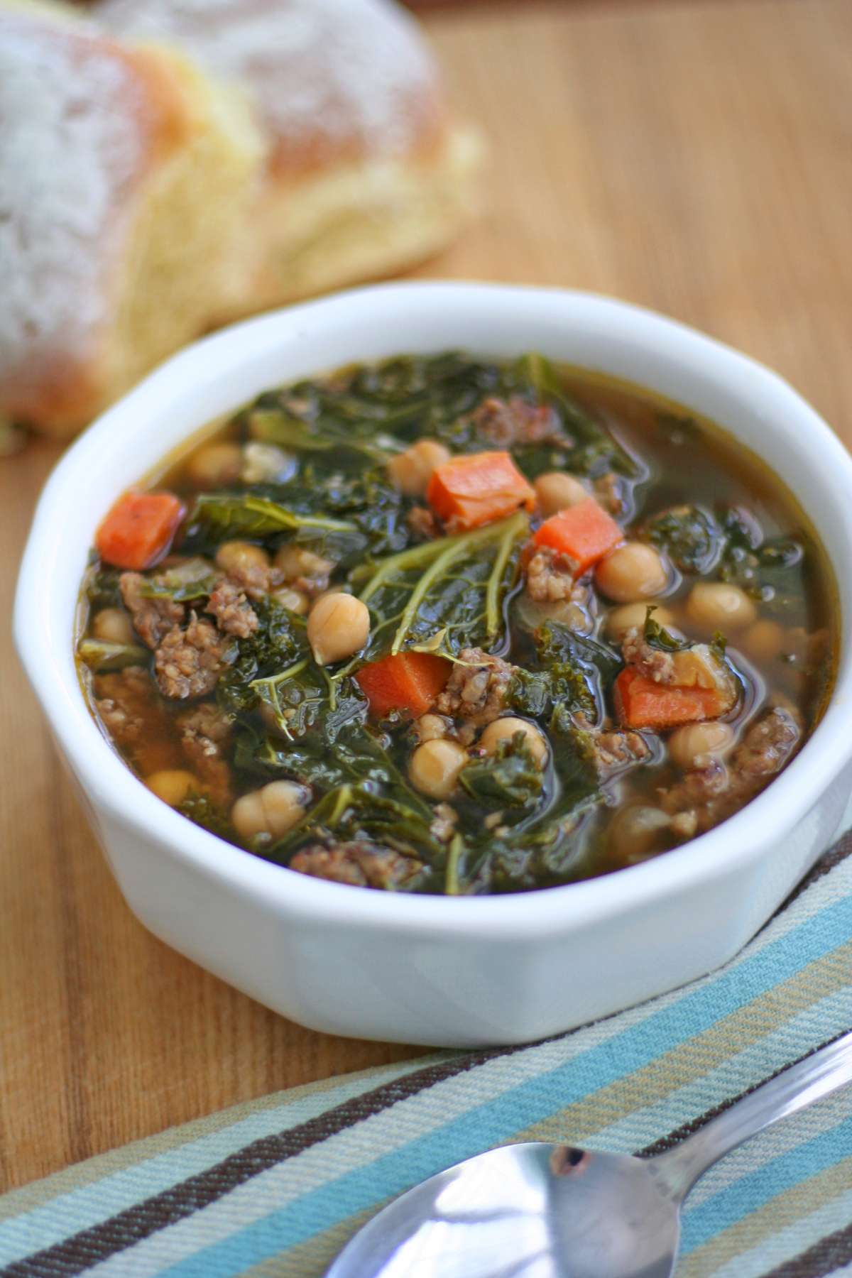 A bowl of Sausage and Kale Soup filled with kale, chickpeas, carrots, and crumbled sausage in a savory broth, served in a white bowl on a wooden table.