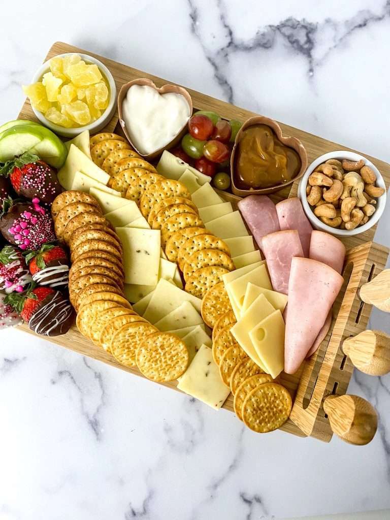 A wooden platter displays an assortment of crackers, sliced cheeses, deli meats, fresh fruits, cashews, yogurt, peanut butter, and candied fruit.