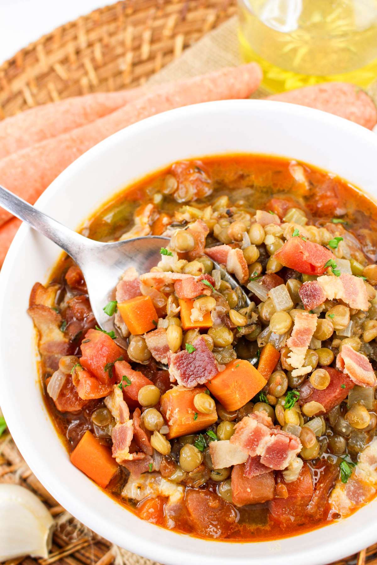 A close-up of a bowl of Bacon Lentil Soup, featuring lentils, diced carrots, tomatoes, and crispy bacon pieces. A spoonful of the soup is lifted, showcasing the hearty mix of ingredients.