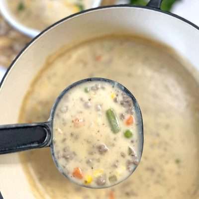 A close-up of a ladle filled with creamy Shepherd’s Pie Soup, featuring ground beef, colorful vegetables, and a thick, savory broth, served in a white Dutch oven.