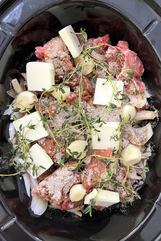 Stew meat in a crockpot with garlic, butter, seasonings, and thyme sprigs.