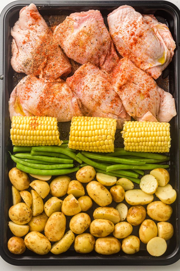 Seasoned chicken thighs, corn on the cob, green beans, and potatoes on a sheet pan.